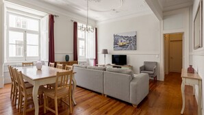 A spacious living room with a large grey sofa, wooden dining table, and chairs. A television is mounted on the wall, and a painting is visible above the sofa. - JOIVY Astonishing 4BR home in Baixa,  5mins to Santa Justa Lift (Lisboa)