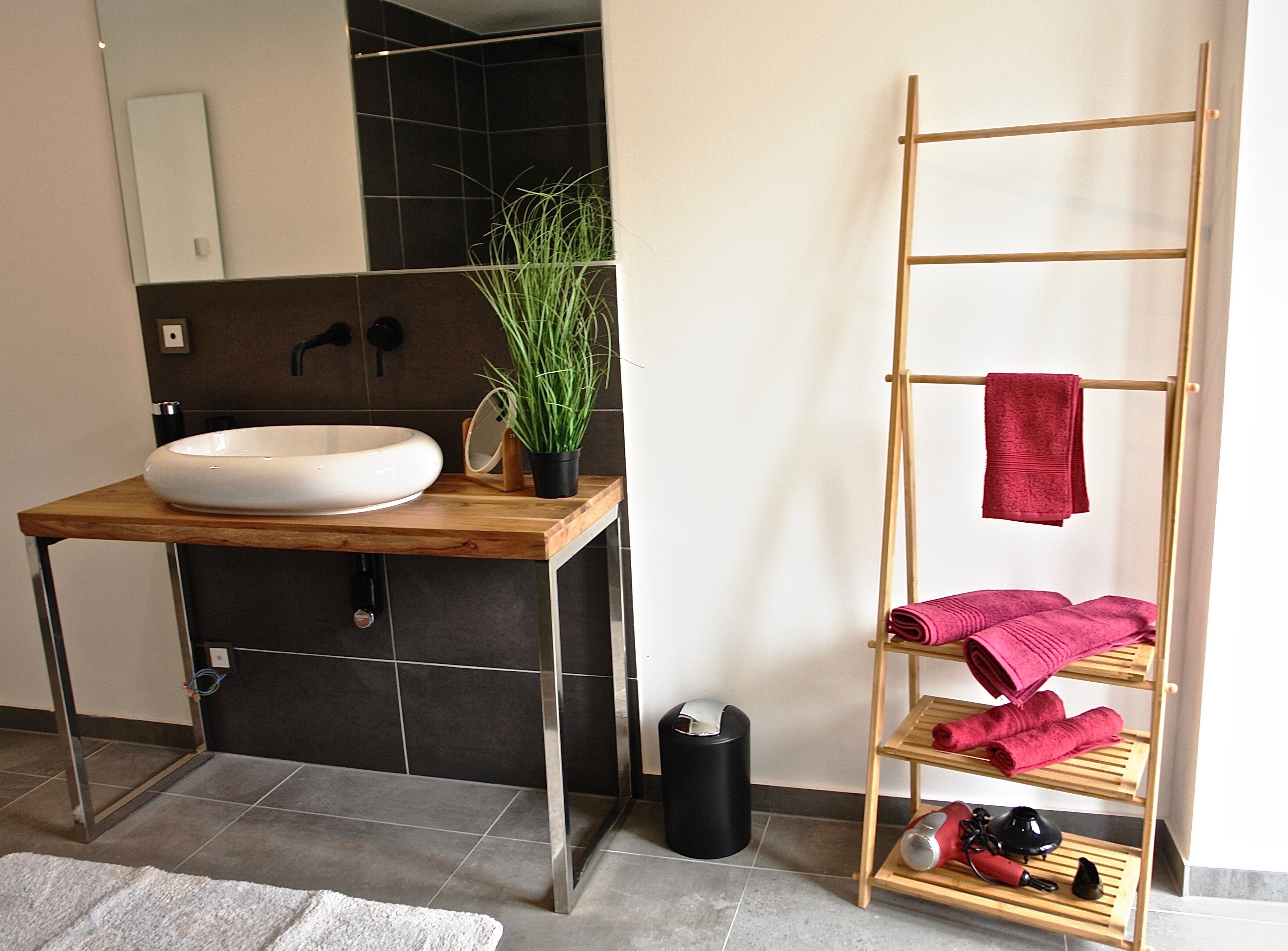 A bathroom with a wooden vanity, a white sink, a mirror, a potted plant, a black trash bin, and a wooden towel rack with towels.