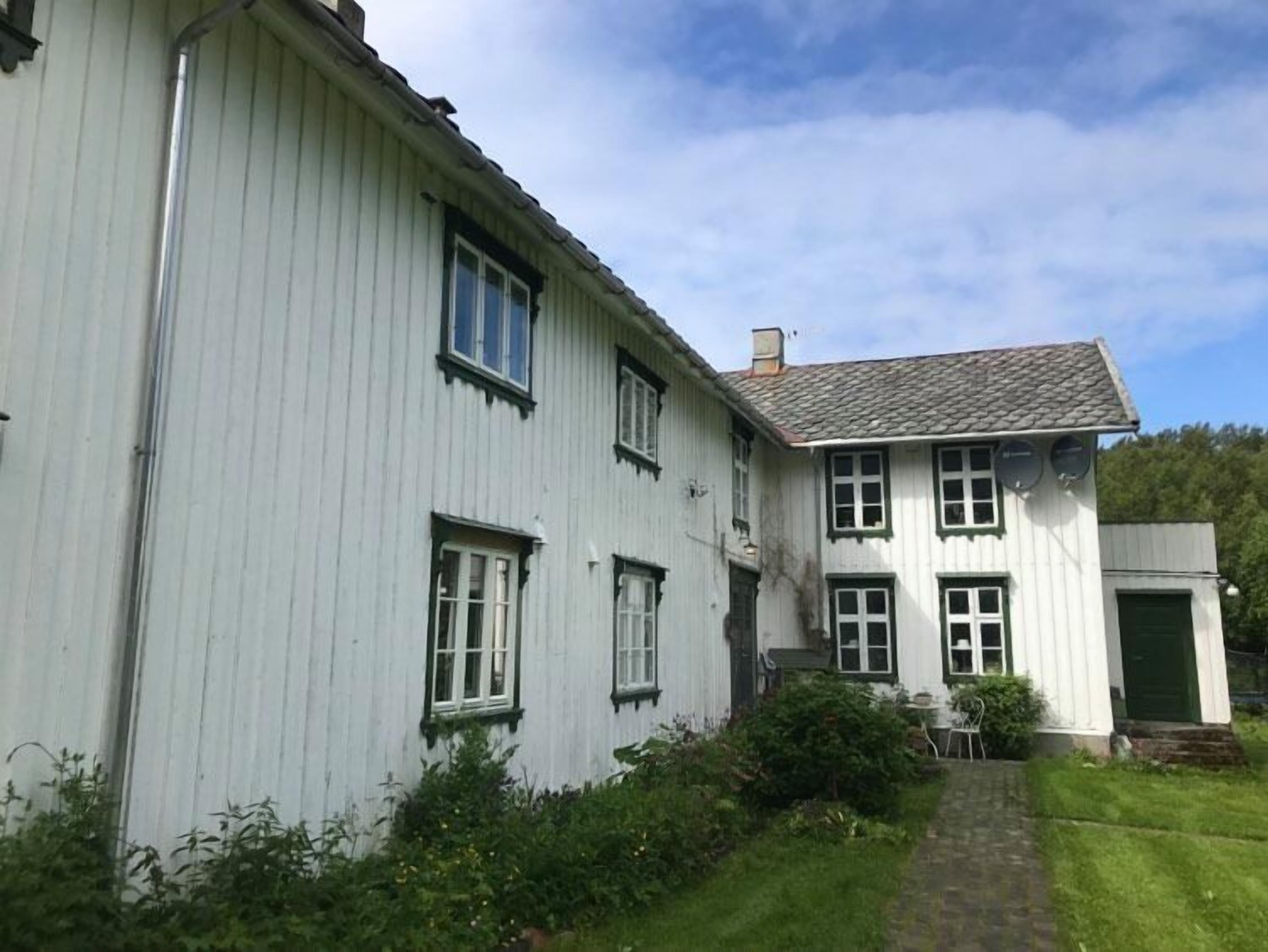Une maison blanche à deux étages, avec une porte verte, un petit porche doté d’une table et de chaises, et un chemin pavé menant à l’entrée.