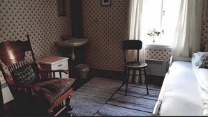 A room with a wooden chair, a small table, a bed, a window with curtains, and a radiator. - Skagen Gaard (Bodo)
