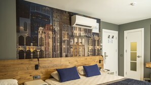 A hotel room with a large cityscape mural on the wall, a wooden headboard, two beds with blue and white bedding, a small bedside table, and a white door. - Pure Blanche Hotel (Antalya)