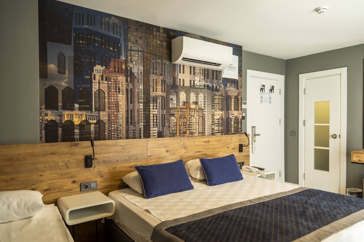 A hotel room with a large cityscape mural on the wall, a wooden headboard, two beds with blue and white bedding, a small bedside table, and a white door.
