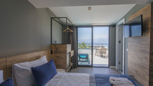 A modern hotel room with a large bed, a wooden headboard, a flat-screen TV, and a sliding glass door leading to a balcony with a view of the mountains. - Pure Blanche Hotel (Antalya)