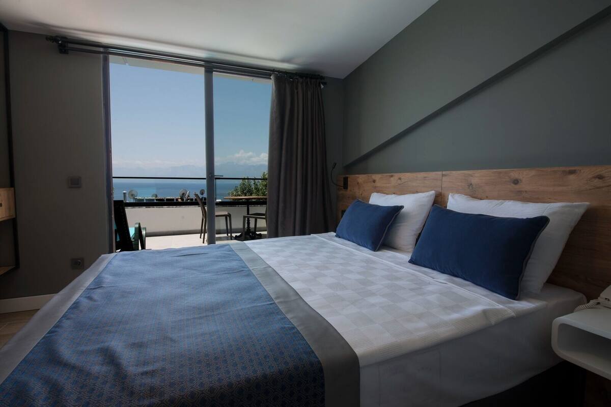 A modern hotel room with a large bed, a wooden headboard, and a balcony with a view of the sea.