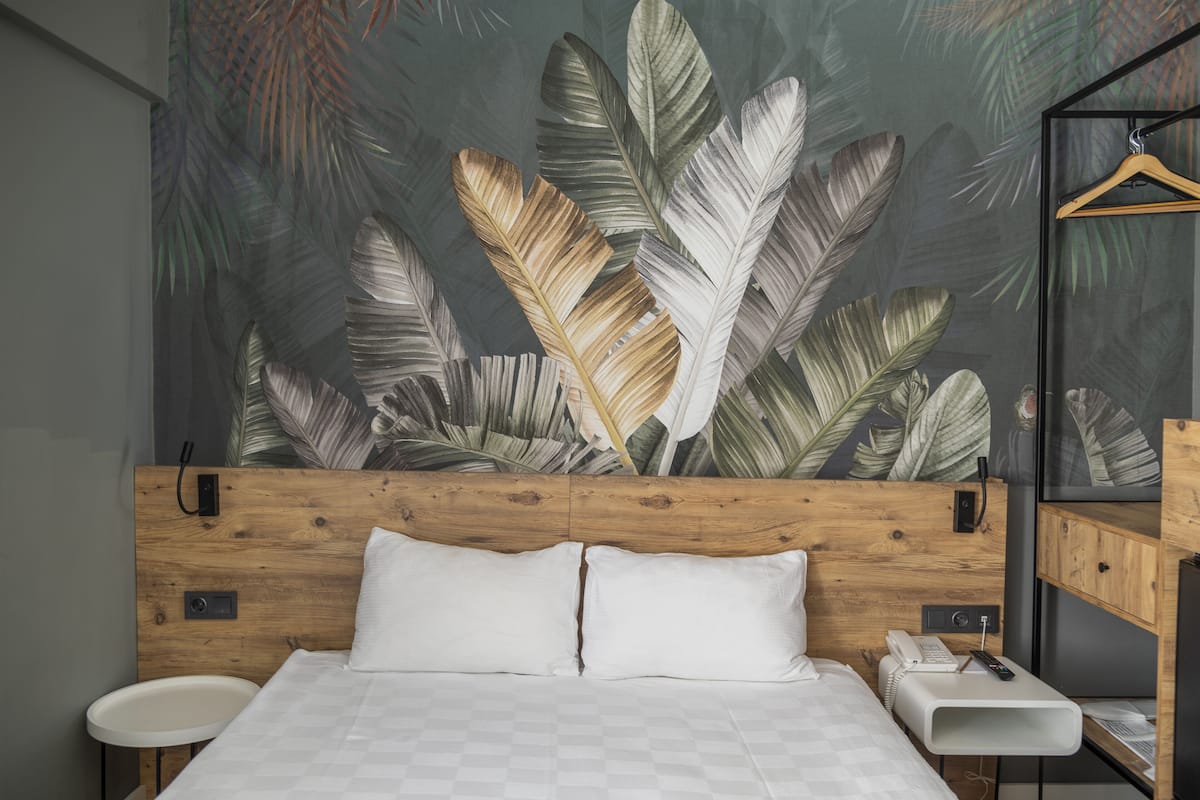 A bedroom with a wooden headboard, a bed with white bedding, a bedside table with a phone, and a wall with a tropical leaf mural.
