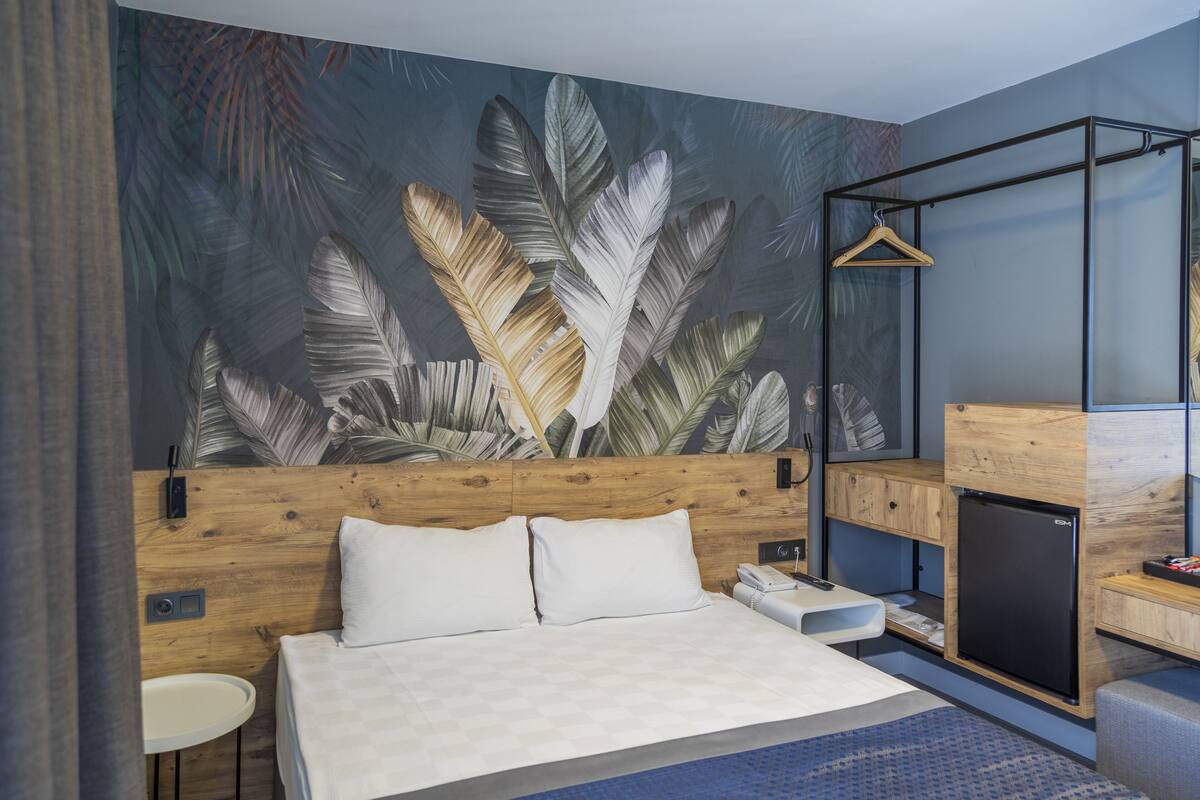 A modern hotel room with a wooden headboard, a bed with white linens, a small bedside table, a compact black refrigerator, and a wall mural of tropical leaves.