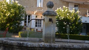 A stone fountain with water spouting from a central column, surrounded by greenery and a building with a tiled roof and white shutters. - La petite Aiguebonne (Allex)