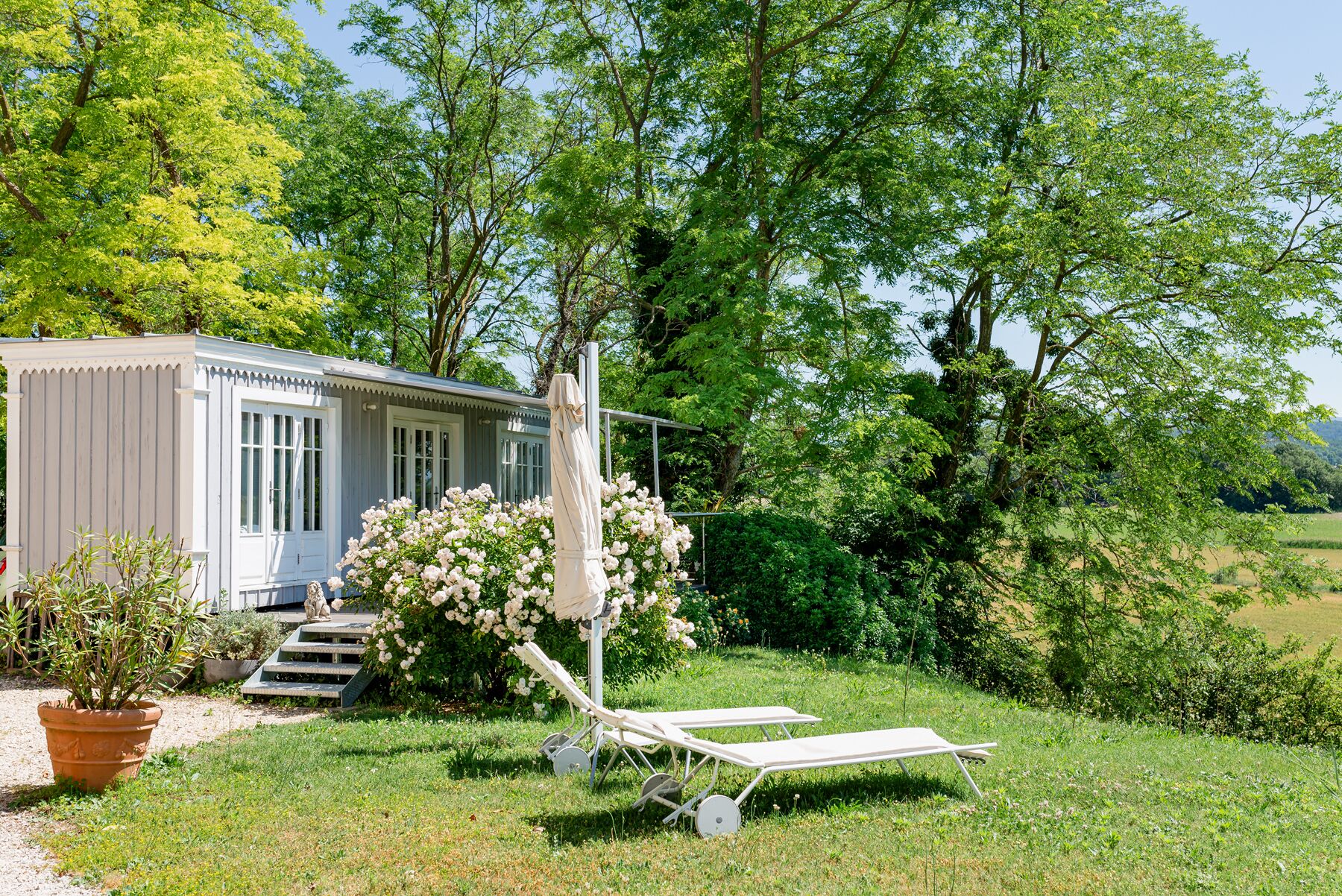 Une petite maison à un seul étage, dotée d’un porche et entourée d’une végétation luxuriante, avec un jardin où poussent des fleurs blanches.