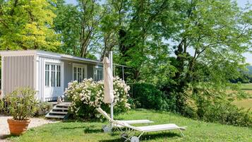 Une petite maison à un seul étage, dotée d’un porche et entourée d’une végétation luxuriante, avec un jardin où poussent des fleurs blanches.