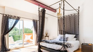 A four-poster canopy bed with a white and patterned bedspread, a small bedside table with a lamp, and a view of a balcony with a chair. - La petite Aiguebonne (Allex)