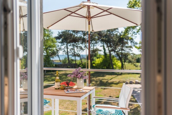 Un balcon avec une table en bois, des chaises blanches, un parasol blanc et une vue sur des arbres et une étendue d’eau.