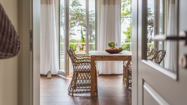 Puerta abierta que da a una habitación con una mesa de madera, sillas de mimbre y vistas a la vegetación exterior.