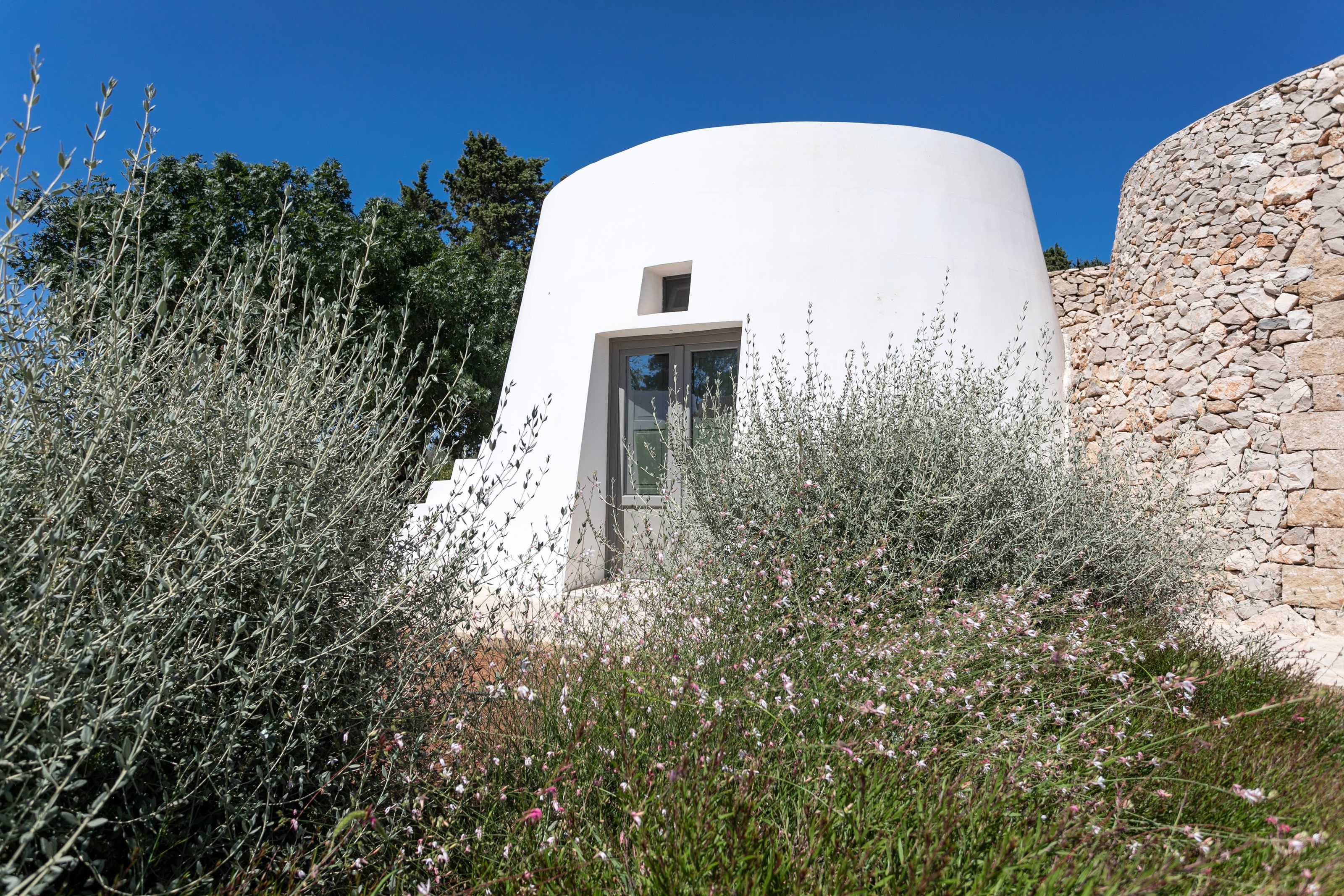 A white building with a small window, surrounded by dense vegetation and a stone wall.