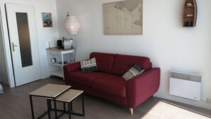 Una sala de estar con un sofá rojo, una mesita, una lámpara, un cuadro enmarcado, una estantería y un radiador.