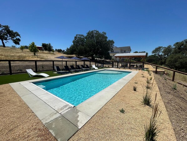 Una piscina rectangular amb una terrassa enrajolada, envoltada per una tanca negra. Hi ha gandules i ombrel·les al voltant de la piscina.