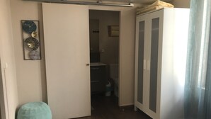 Une chambre à coucher comprenant un lit, une armoire, une porte donnant sur une salle de bain et un petit tabouret.