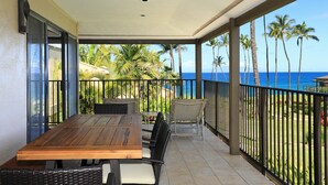 A balcony with a wooden table and chairs, overlooking a tropical garden and the ocean. - Wailea Elua 1602: Oceanview Oasis W/ Easy Beach Access Amidst Tropical Splendor (Wailea, Maui)