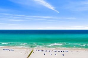 A beach with rows of blue umbrellas and lounge chairs, a sandy shore, and a clear blue sky.