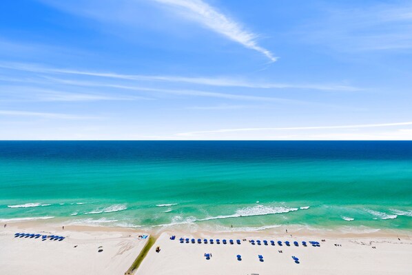 A beach with rows of blue umbrellas and lounge chairs, a sandy shore, and a clear blue sky.