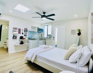 Ein modernes Schlafzimmer mit Bett, Deckenventilator, einer kleinen Küchenzeile und einem durchgegangenen Tür erkennbaren Badezimmer.
