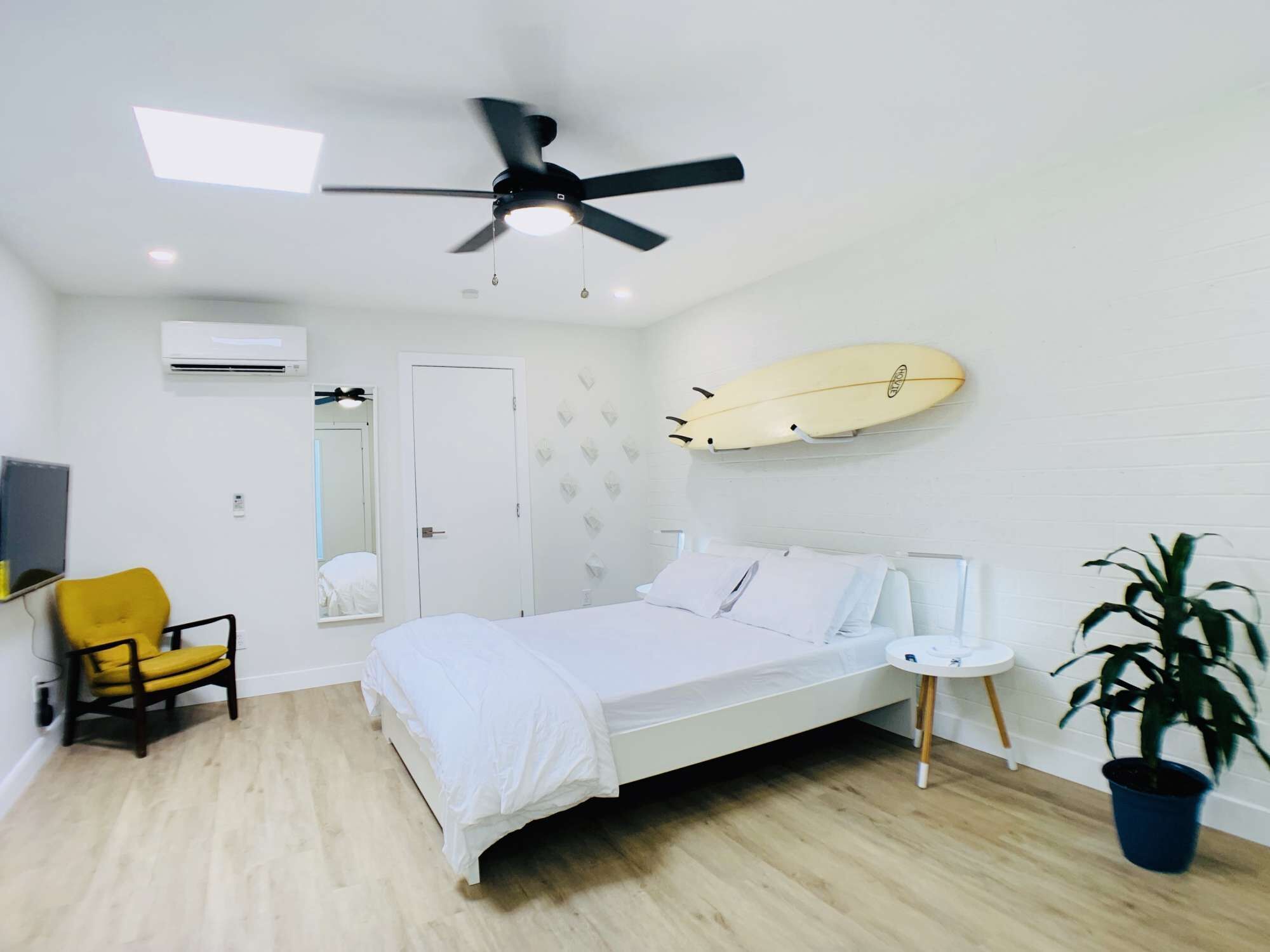 Een moderne slaapkamer met een bed, een plafondventilator, een surfboard aan de muur, een stoel, een klein tafeltje en een potplant.