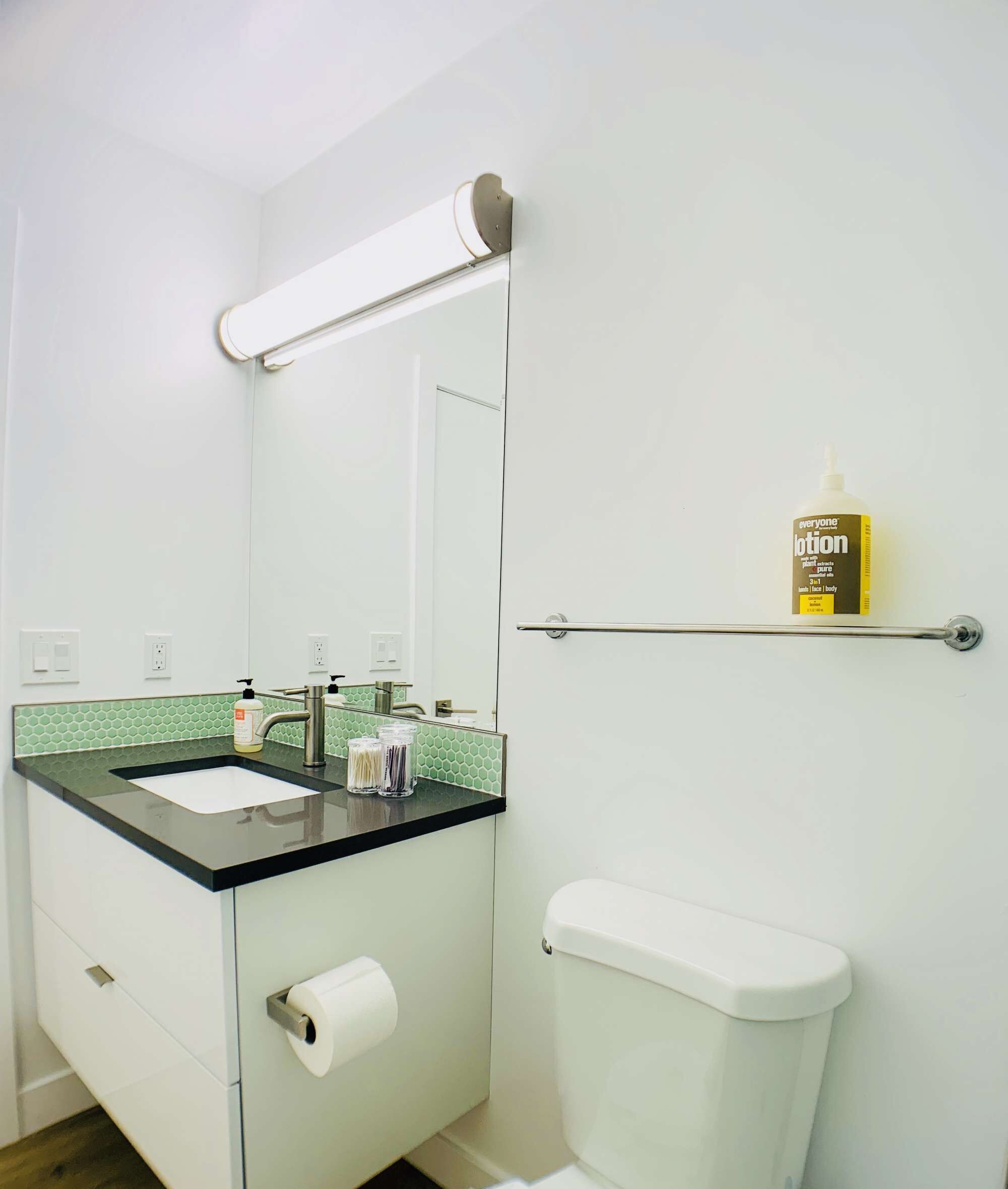 A bathroom with a white toilet, a sink with a mirror above it, and a shelf with a bottle of lotion.