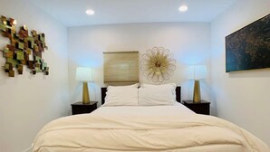Quarto com uma cama grande, duas mesas de cabeceira com abajures, um detalhe decorativo na parede e um quadro emoldurado.