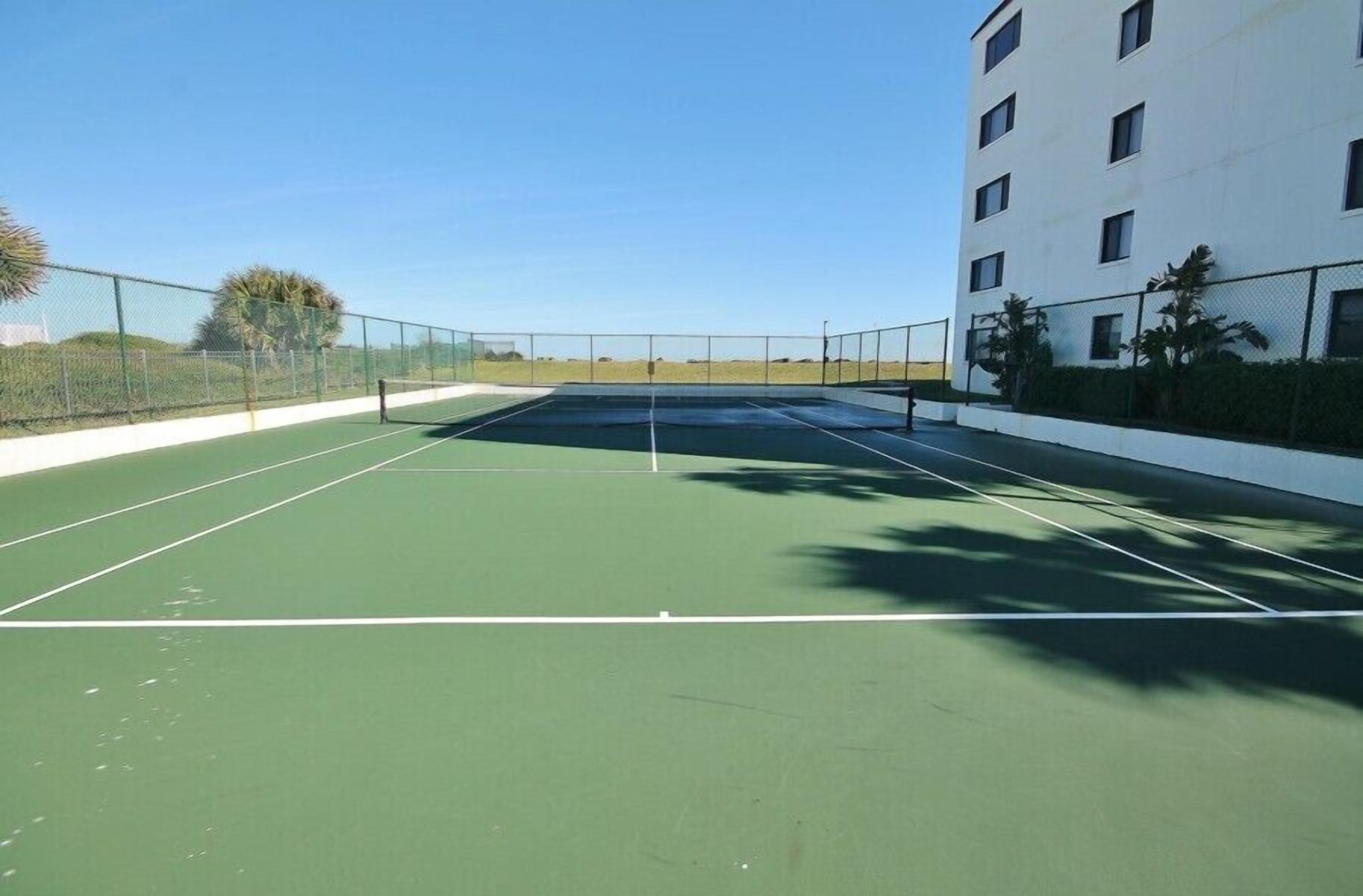 An outdoor tennis court with a clear sky, surrounded by a white building and a fence.
