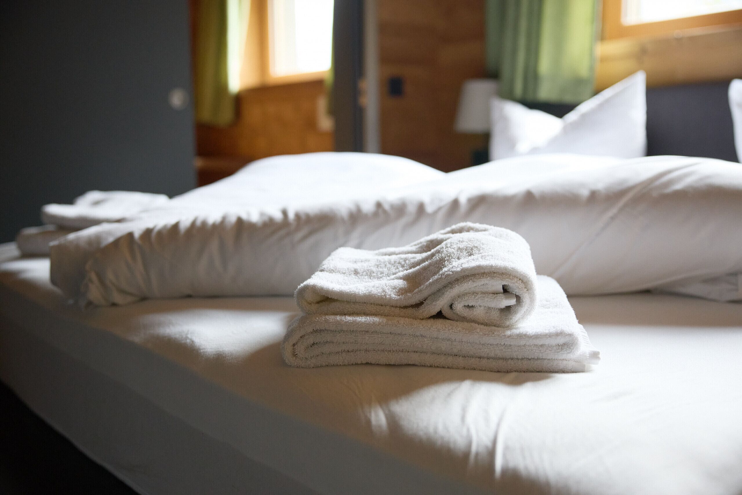 Un letto rifatto con cura, lenzuola e cuscini bianchi, e un asciugamano piegato sul letto. Si vede una finestra con tende sullo sfondo.