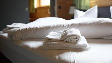 Un letto rifatto con cura, lenzuola e cuscini bianchi, e un asciugamano piegato sul letto. Si vede una finestra con tende sullo sfondo.