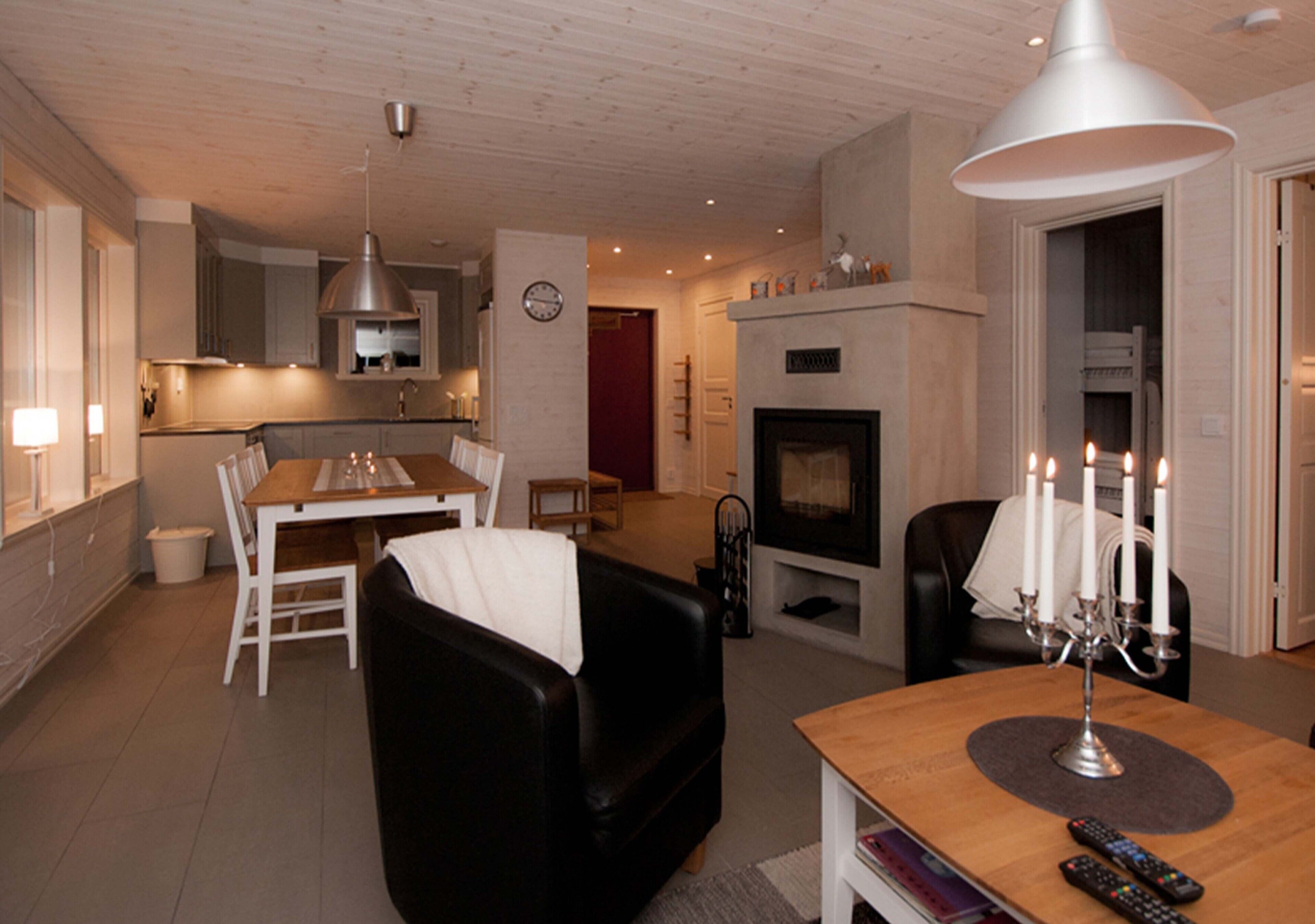 Un salon moderne avec une cheminée, un coin repas avec une table et des chaises en bois, et une cuisine en arrière-plan.