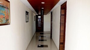 A long, narrow hallway with white walls and a wooden ceiling. The floor has a pattern of white and dark tiles. - The New Holiday II (Mahesana)