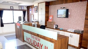 A reception desk labeled "The New Holiday II" with a television, wooden cabinets, and a calendar on the wall. - The New Holiday II (Mahesana)