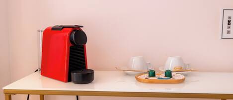 A red coffee machine on a white table with two white cups and saucers, a tray with a green candle, and a power outlet.