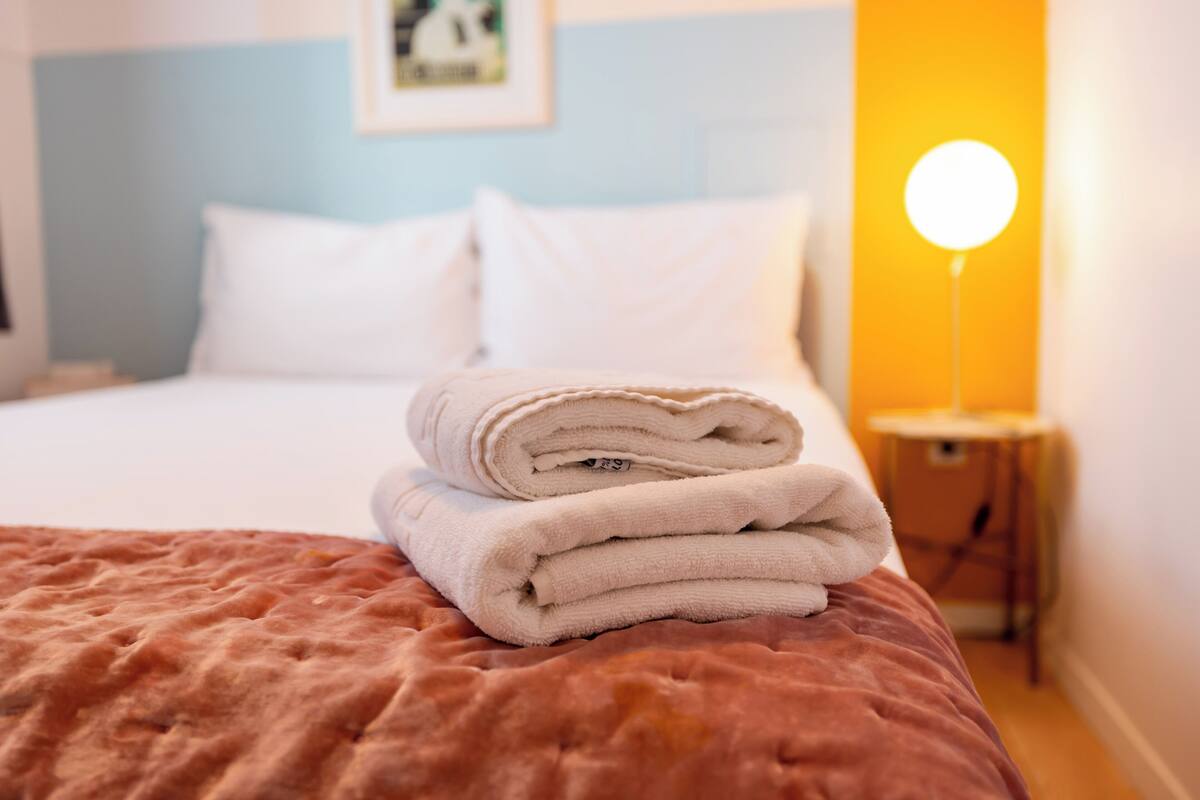 A neatly made bed with a folded white towel, a bedside lamp, and a wall-mounted picture.