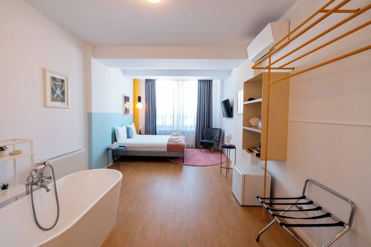 A modern hotel room with a large bathtub, a bed with a view, a desk, and a TV.