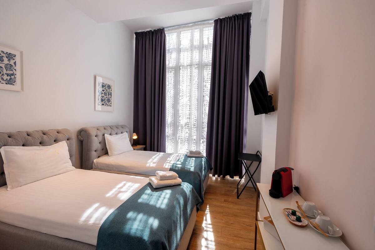 A hotel room with two beds, a desk, a chair, a coat rack, and a window with curtains.