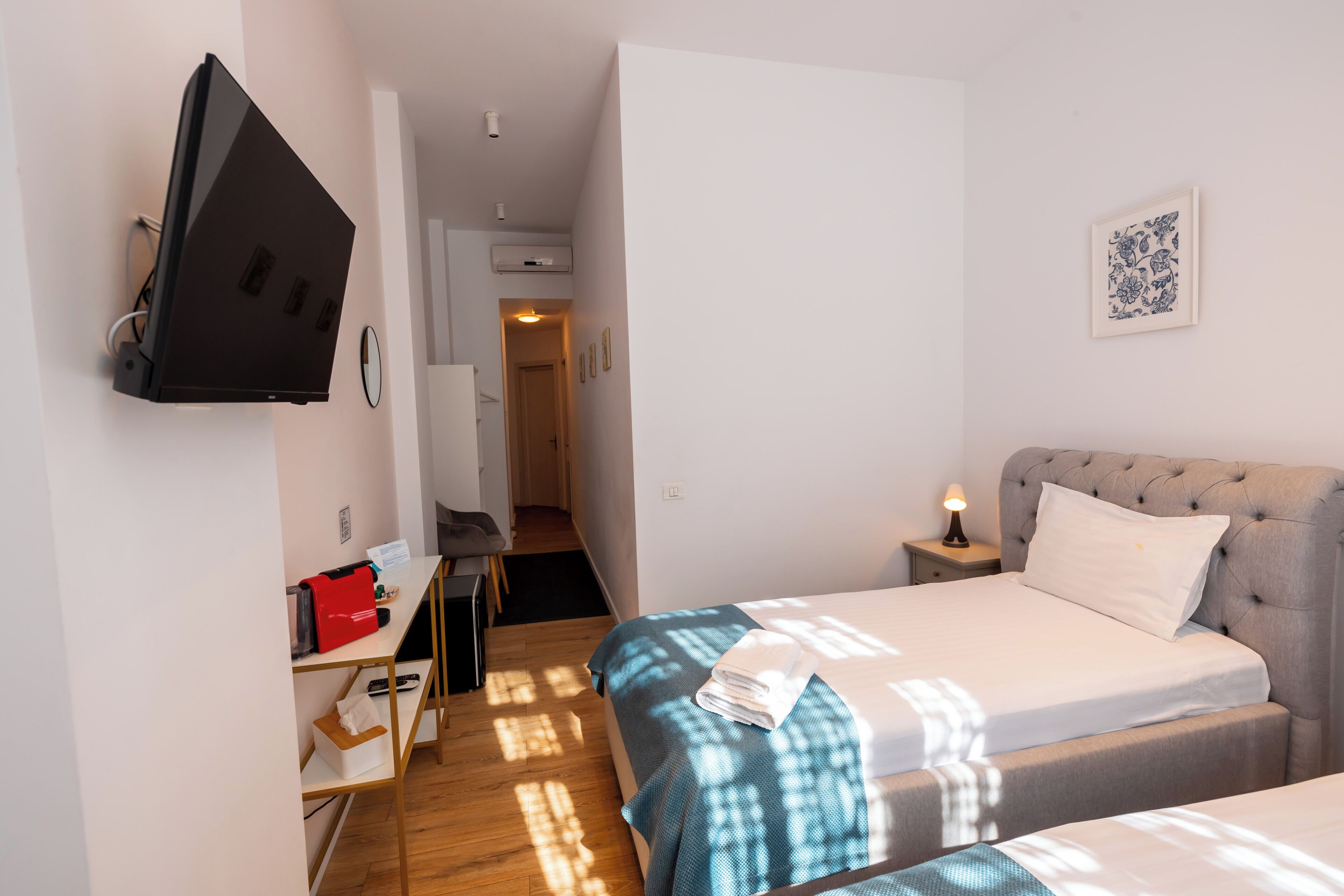 Habitación de hotel con cama, televisor montado en la pared, escritorio con silla y una mesita de noche con lámpara.