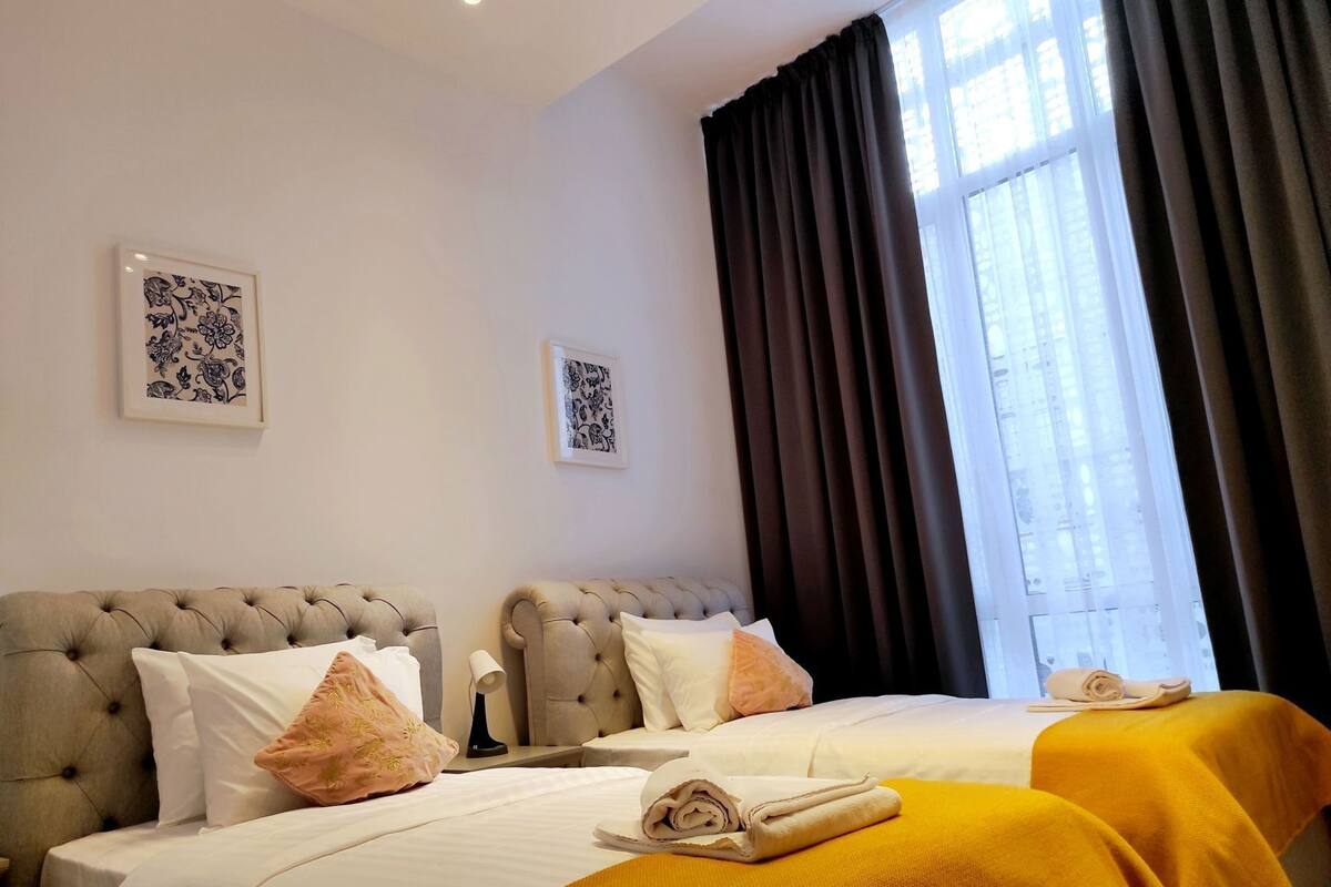 A hotel room with two beds, a large window with curtains, and two framed artworks on the wall.