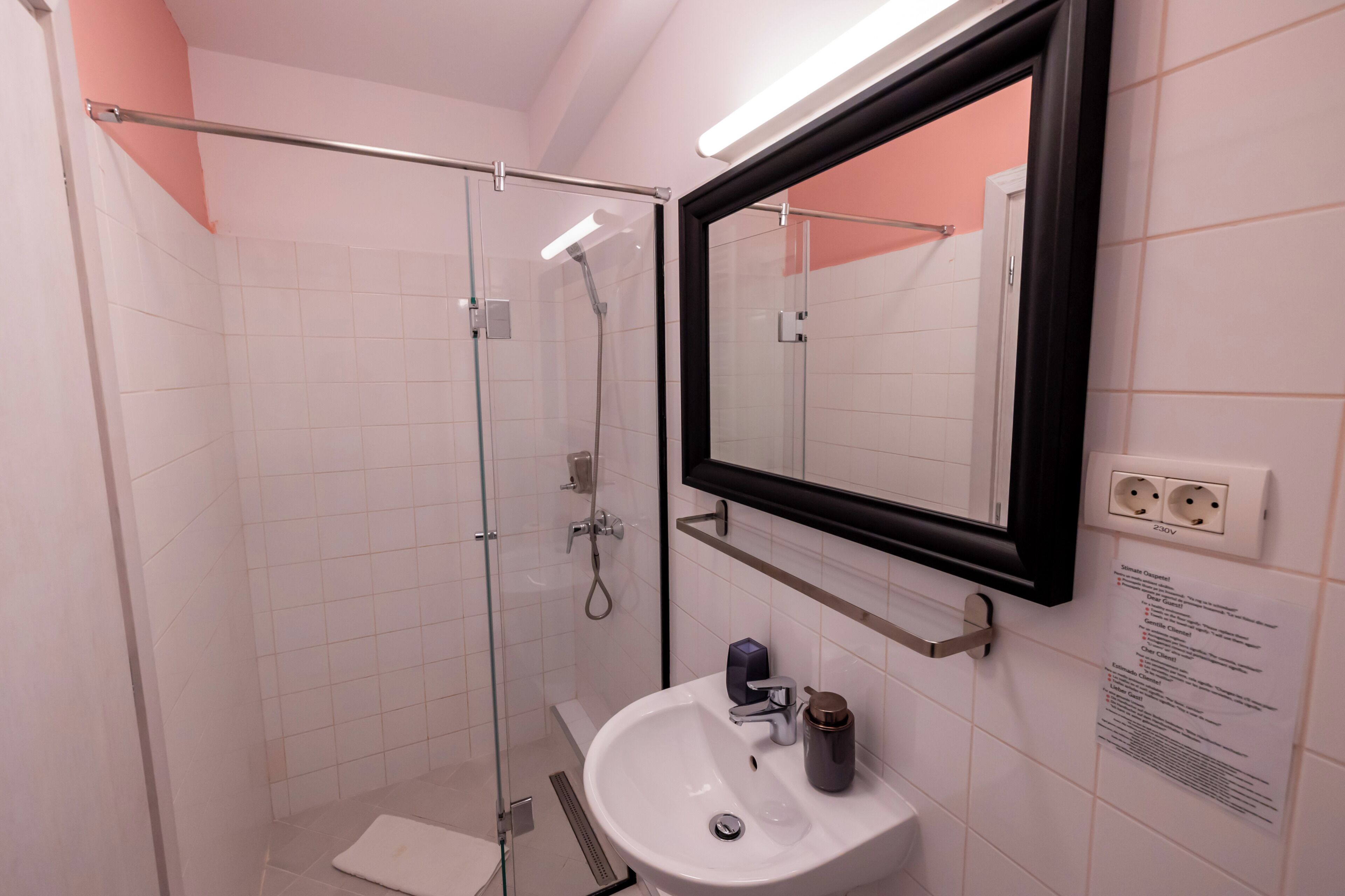 A modern bathroom with a glass shower enclosure, a white sink, a mirror, and a wall-mounted electrical outlet.