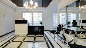 En modern hotellobby med ett svart- och vitrutigt golv, en kristallkrona, en receptionsdisk och stora fönster med persienner.