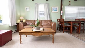 A living room with a brown leather sofa, a wooden coffee table with a plant, a red ottoman, a dining table with chairs, and a window with curtains.