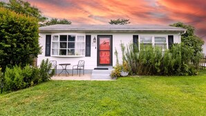 A single-story house with a red door, white siding, and black shutters. There is a small front porch with two chairs and a table.