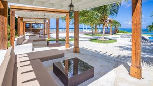 Un espace extérieur aménagé, abrité et doté de bancs rembourrés et d’une table à plateau en verre, entouré de piliers en bois et de palmiers, avec vue sur une plage.