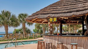 Un bar junto a la piscina con taburetes de madera, techo de paja y palmeras al fondo.
