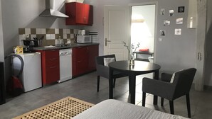 Une cuisine compacte avec des placards rouges, un four à micro-ondes et une machine à café. On y trouve un coin repas avec une table et des chaises, ainsi qu'un lit recouvert d'une courtepointe à motifs.