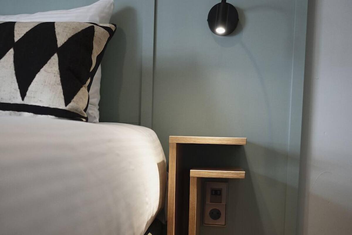A neatly made bed with a patterned pillow, a bedside table with a lamp, and a wall-mounted switch.