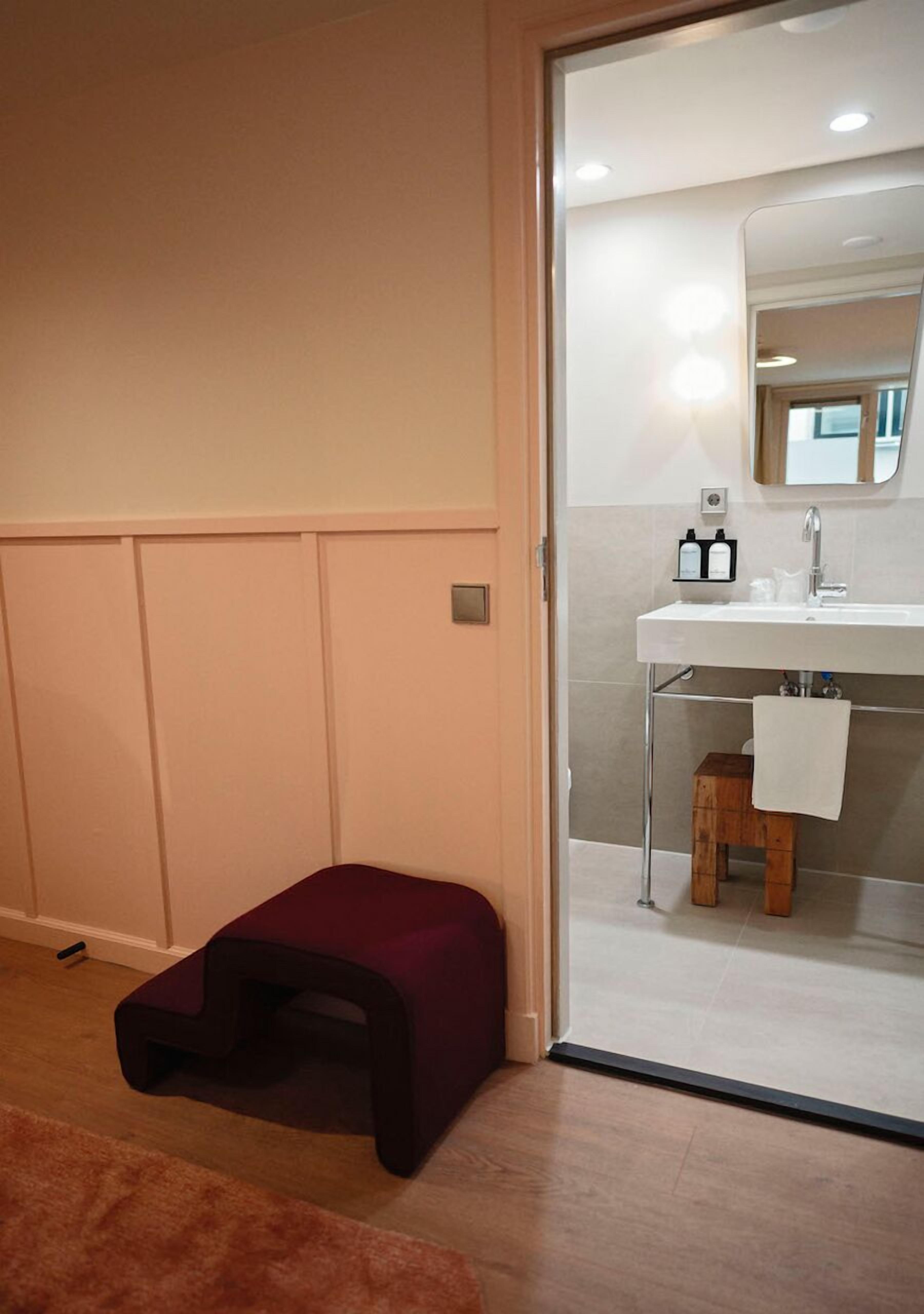 A room with a wooden floor, a small wooden stool, a purple bench, and a bathroom with a white sink and mirror.