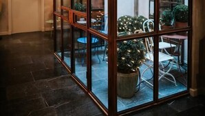 Ein durch eine Glasscheibe abgetrennter Raum mit Blick auf einen Außenbereich mit Tischen, Stühlen und einem Blumentopf.