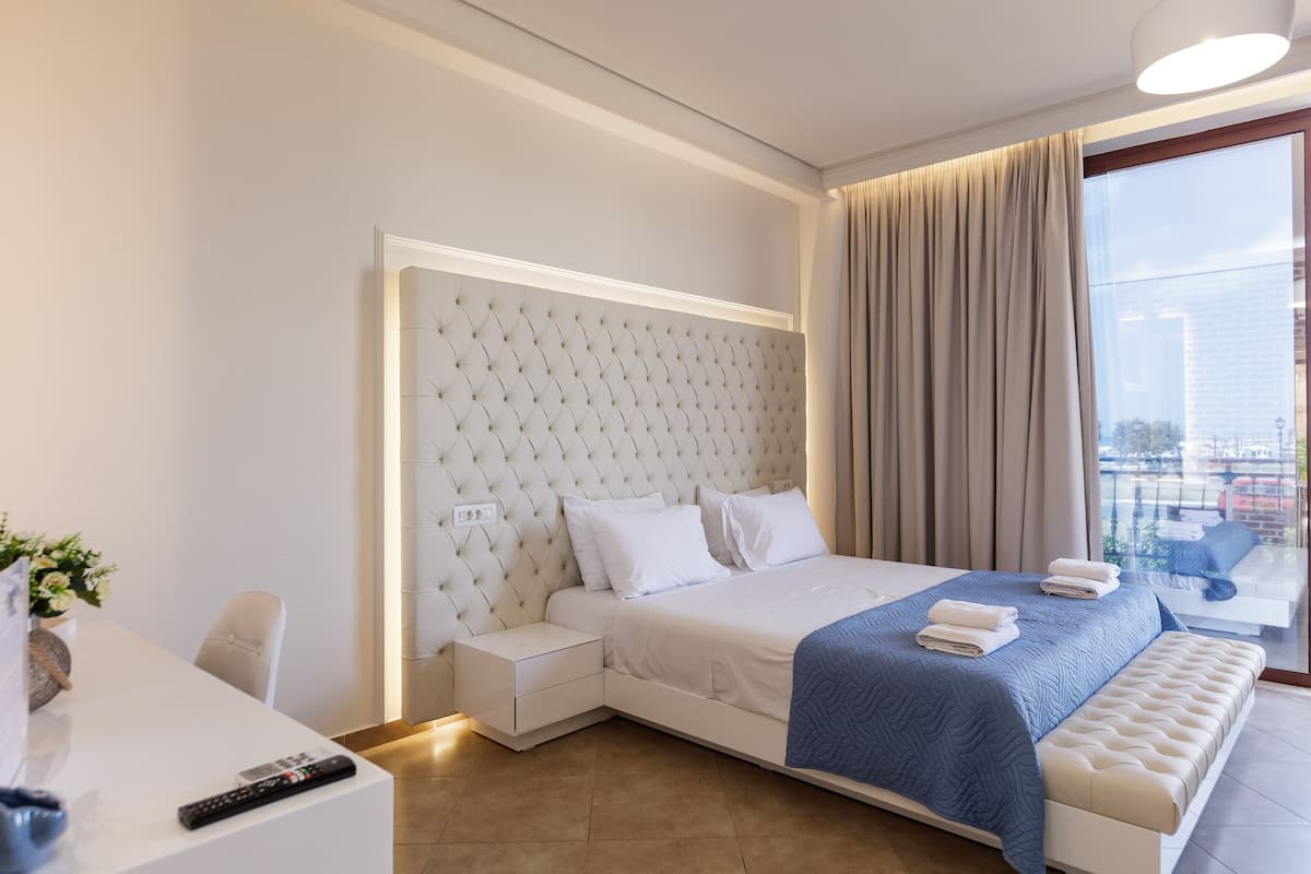 A modern hotel room with a large bed, a bedside table, a desk, and a view of the city through a large window.